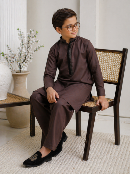 Boy wearing a brown traditional outfit sitting on a wooden chair indoors.