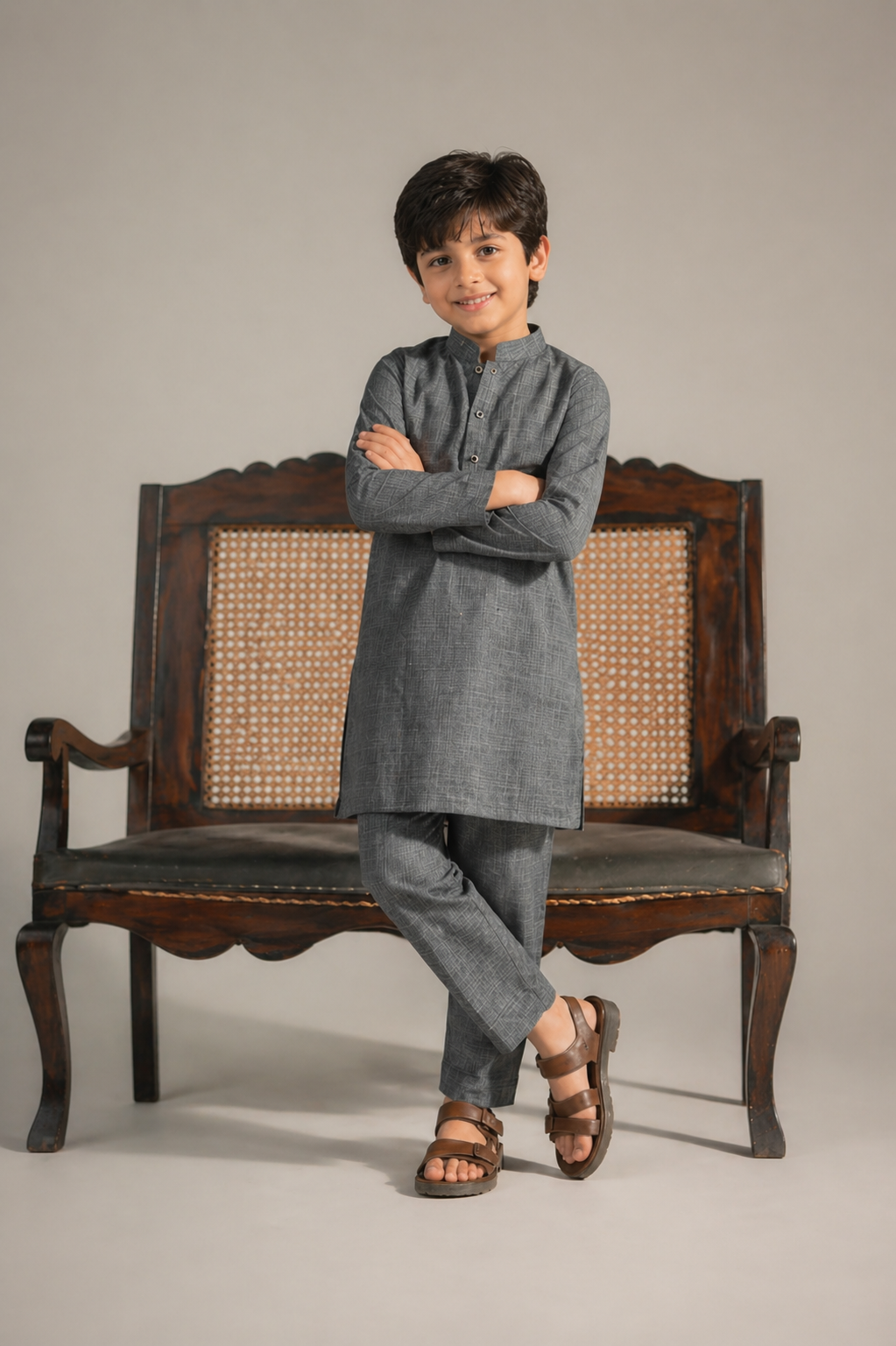 Child wearing a gray kurta with crossed arms on a wooden chair against a plain background
