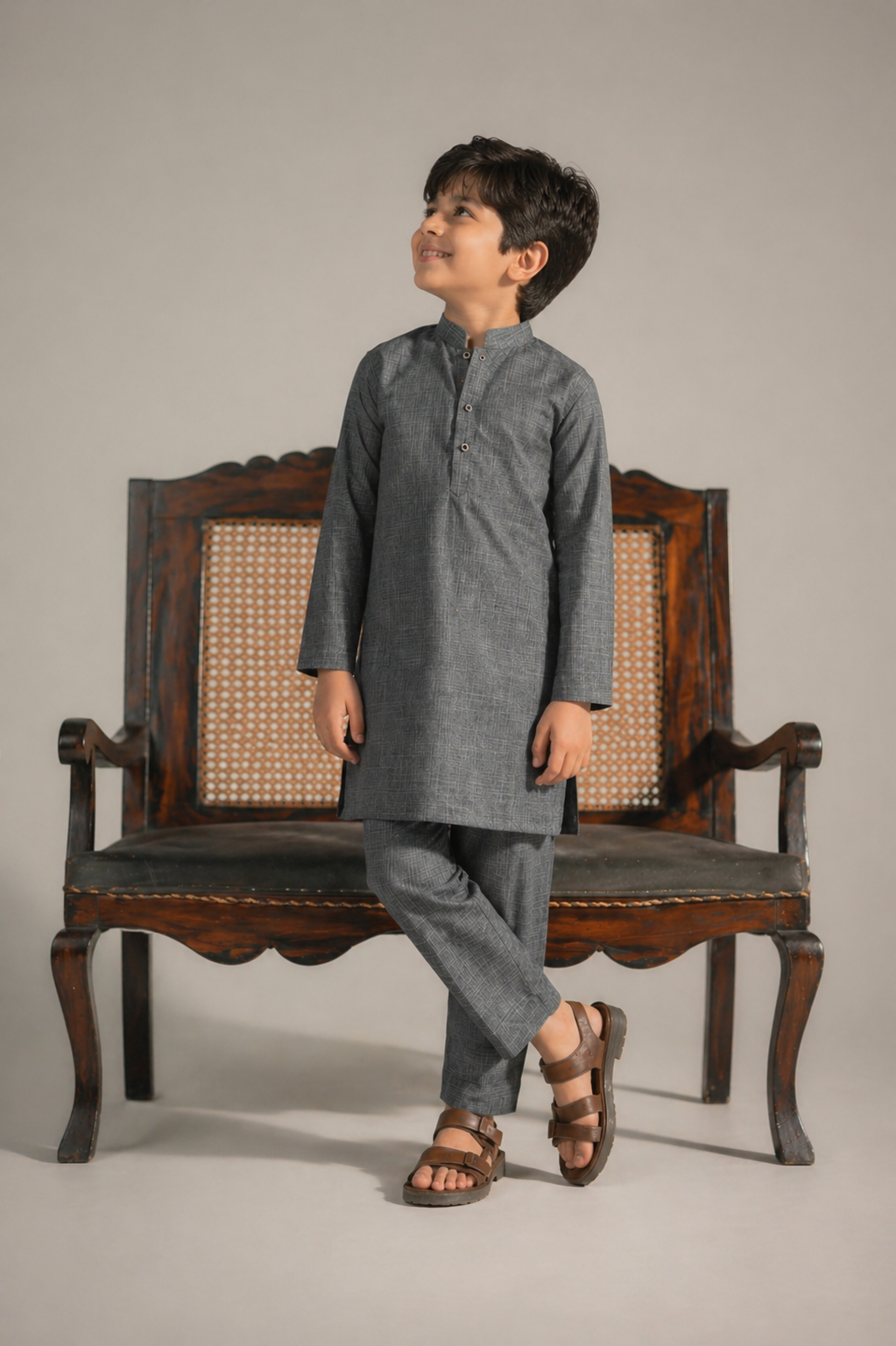 Child wearing a gray kurta standing next to an ornate wooden chair on a plain background