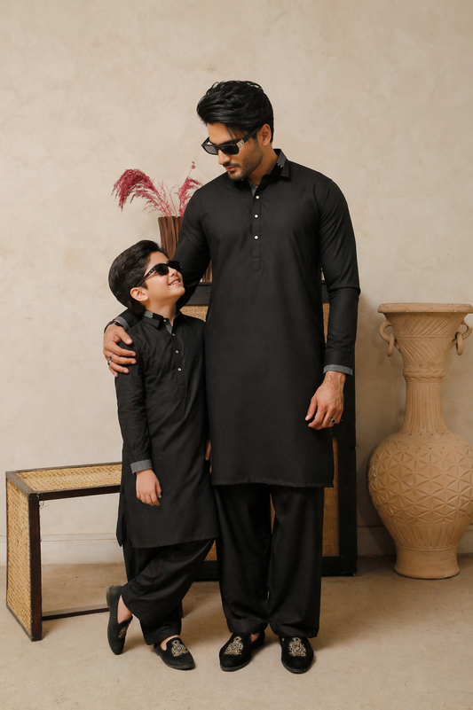 Father And Son in black traditional outfits standing close together against a beige wall.