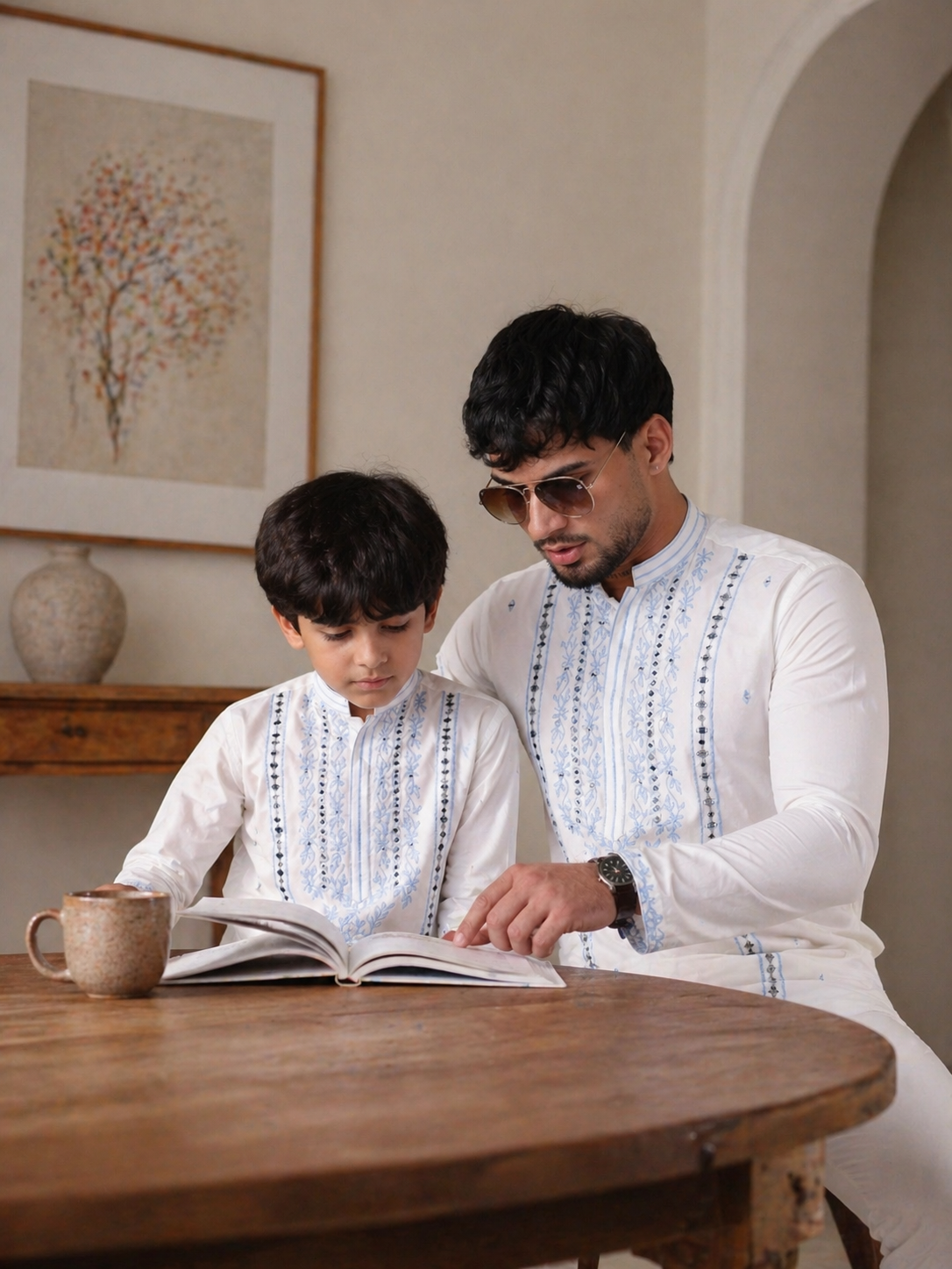 Father & Son in traditional attire sitting at a table with an open book, in a room with a decorative wall piece.