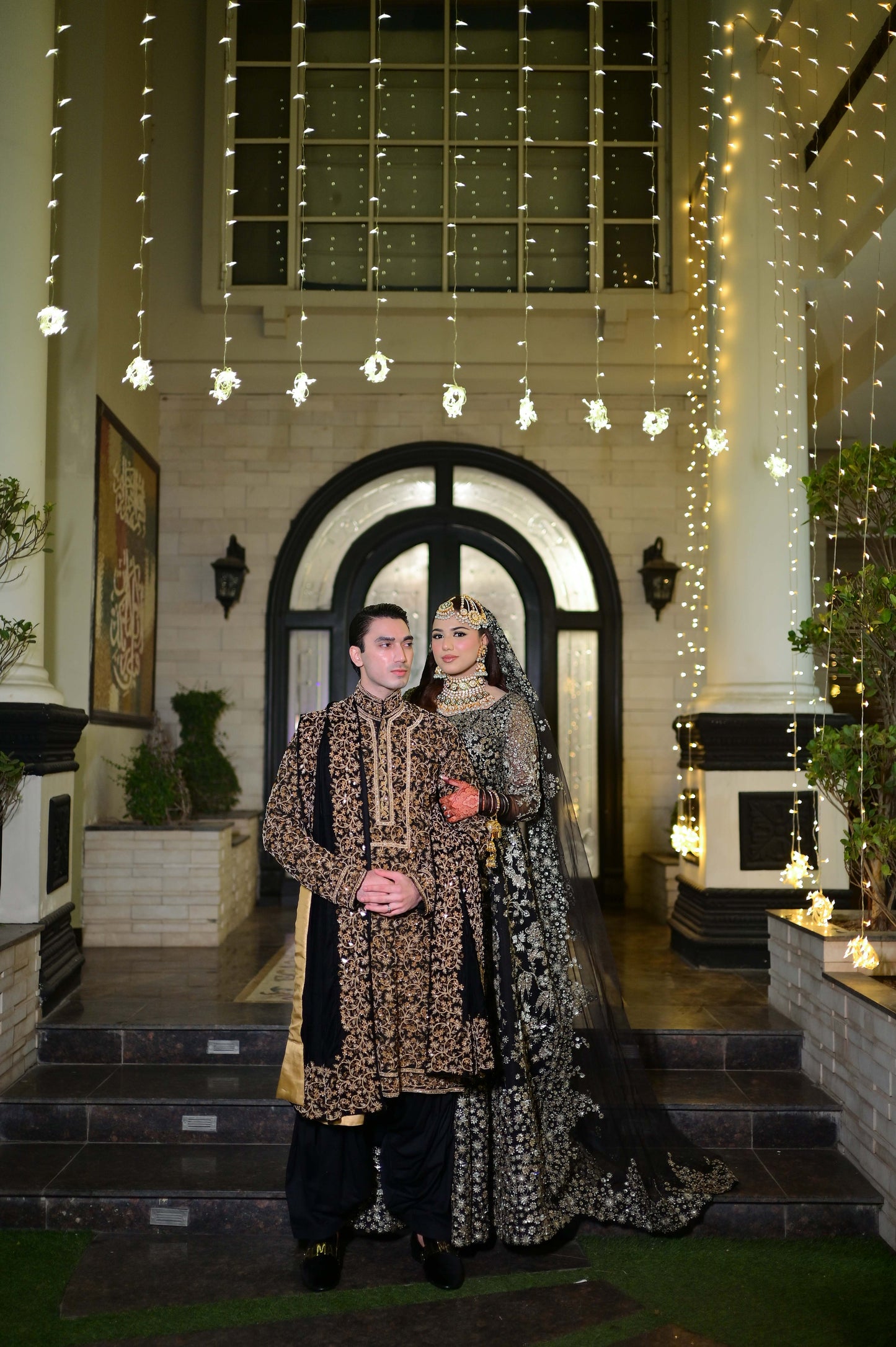 Two people in traditional attire standing in a decorated indoor setting with string lights.