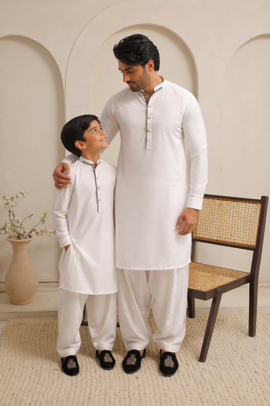 Man and child in matching white outfits standing in a room with a decorative wall and chair.