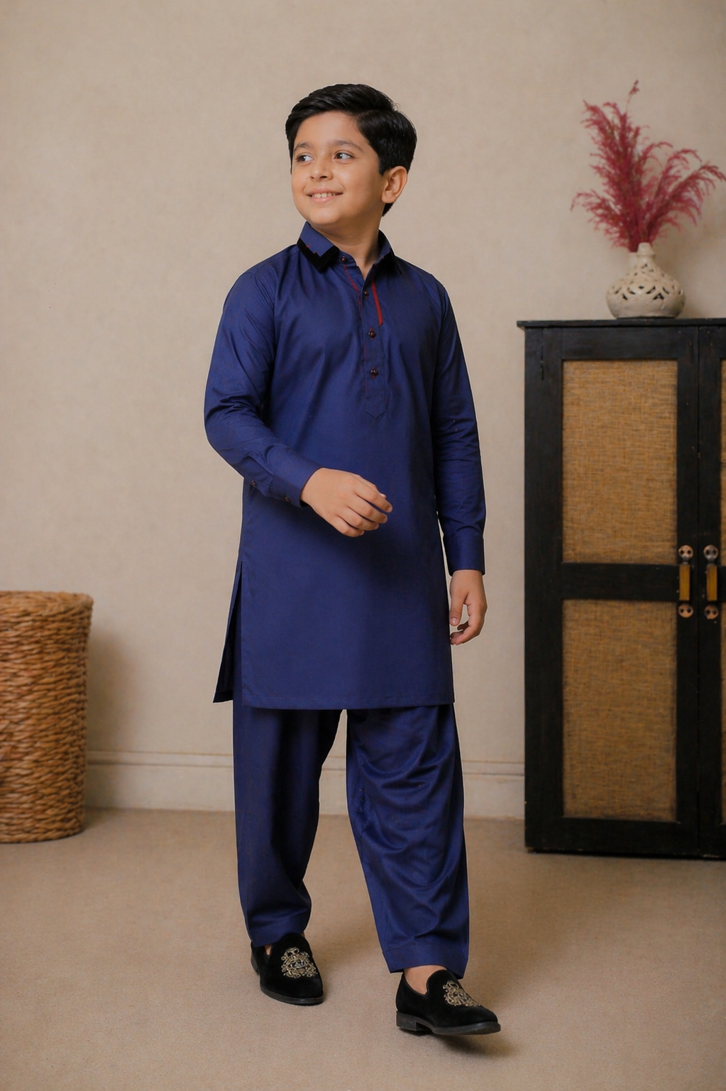 Young boy wearing a blue traditional outfit standing in a room with a beige wall and wooden cabinet.