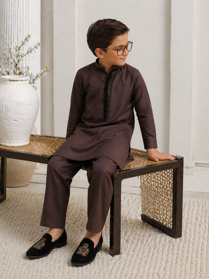 Young boy in a brown traditional outfit sitting on a woven bench.