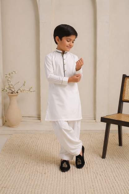 Young boy in a white traditional outfit standing in a room with a chair and vase in the background.