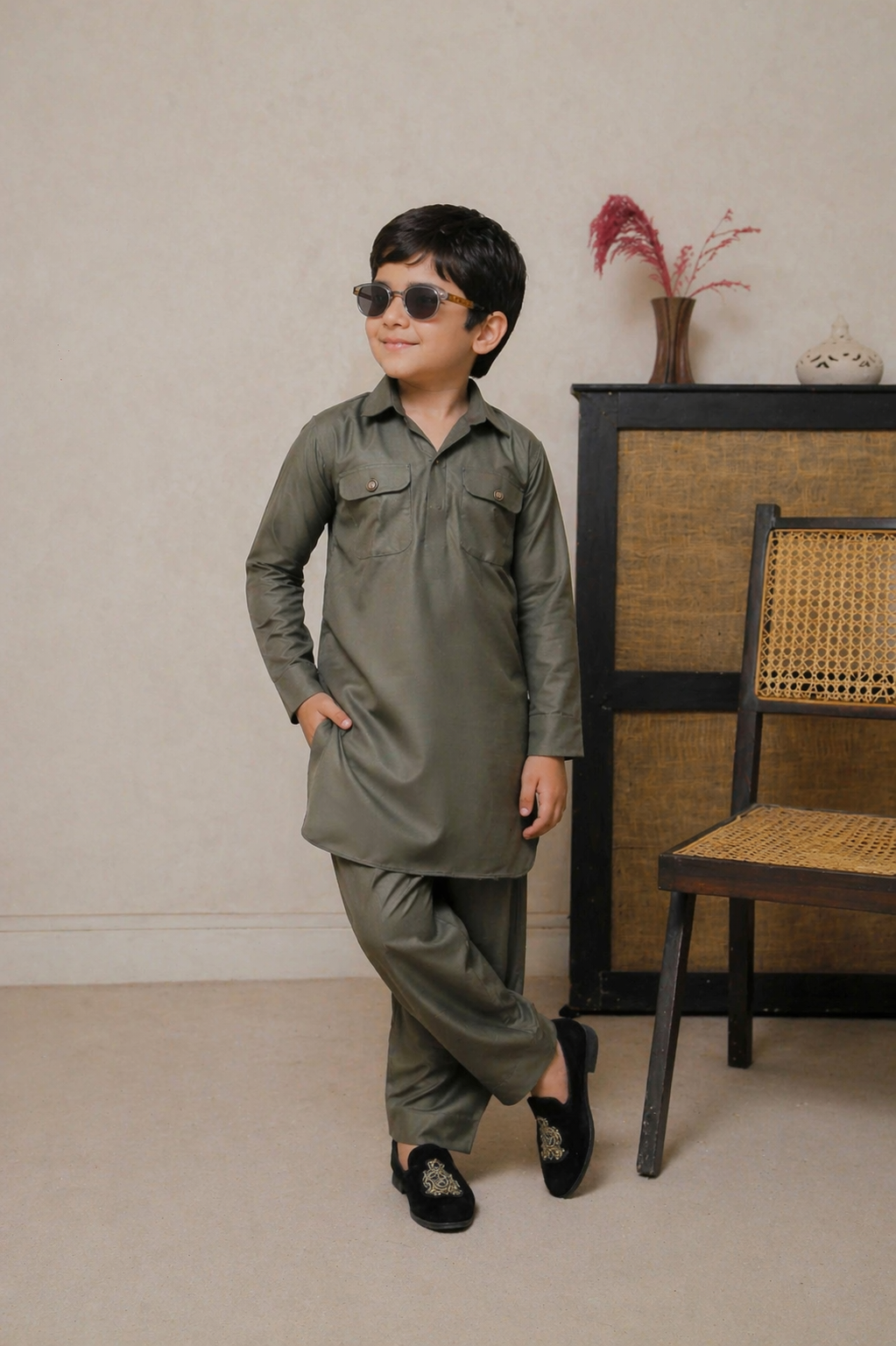 Child wearing a green outfit with sunglasses standing in front of a beige wall with a chair and decorative items.