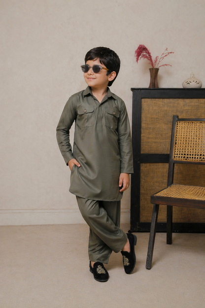 Child wearing a green outfit with sunglasses standing in front of a beige wall with a chair and decorative items.