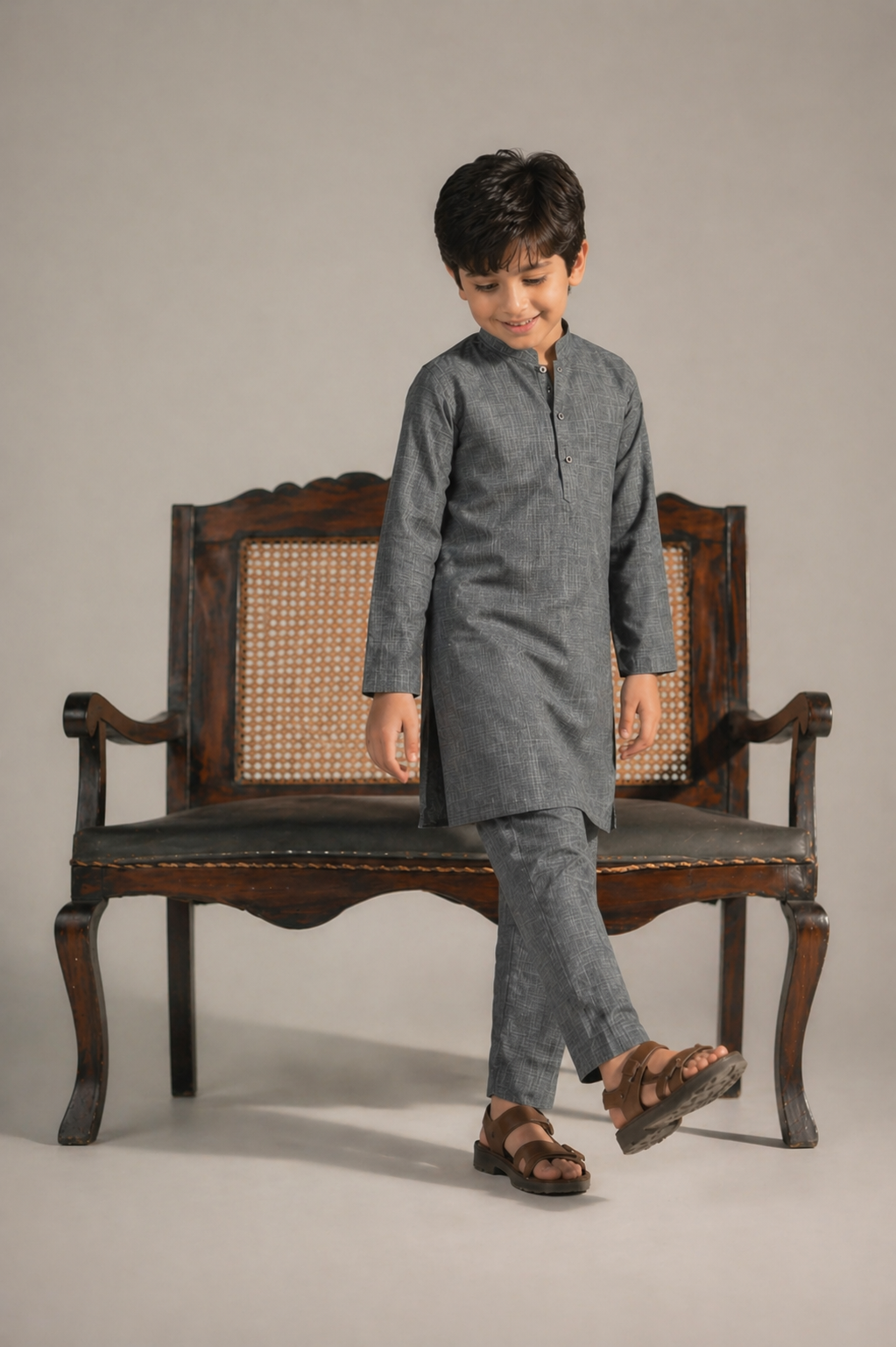 Child wearing a gray kurta standing next to an ornate wooden chair on a plain background